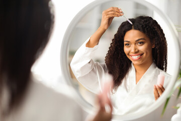 Smiling Black Woman Applying Serum For Hair Repair While Standing Near Mirror - obrazy, fototapety, plakaty