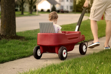 Beautiful young girl sitting in a red wagon cart by the road outdoors. Father is pulling a wagon with daugther.