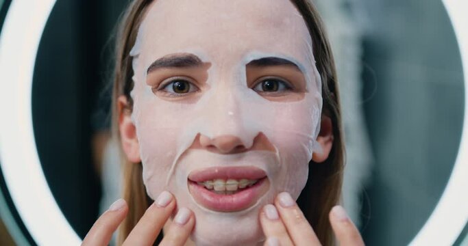 Close up of young girl with facial mask looking in camera on background of the mirror in bathroom. Cosmetic procedure. Beauty spa and cosmetology