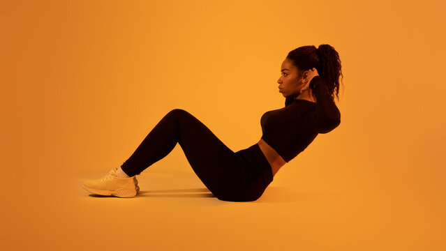 Black fit woman doing v-ups abdominal exercise, sitting on floor on orange neon studio background, side view