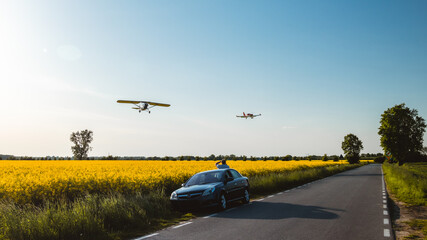 Airplane flying over field of rape. Man taking picture. Car in the middle. Two planes.