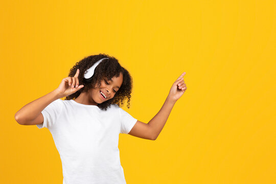 Glad curly teen african american girl in white t-shirt and wireless headphones dancing, listen music