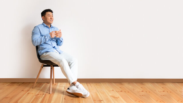 Cheerful Asian Middle Aged Man Sitting On Chair At Home, Drinking Herbal Tea Or Coffee, Looking At Copy Space, Panorama