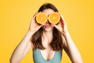 Playful lady in swimsuit holding orange circles over her eyes and having fun on yellow studio background