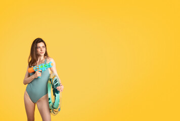 Cheerful woman in swim clothes posing with water gun and inflatable ring, having fun on yellow background, free space