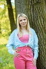 Amazing young blonde in pink skinny jeans and blue shirt standing near tree. Blue forget-me-nots in pockets of jeans.