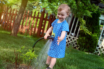 Adorable little girl watering  tree  with a garden hose on hot and sunny summer evening