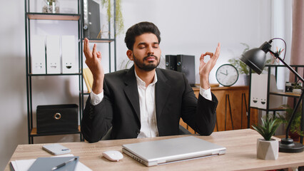 Indian businessman working on laptop computer, meditating, doing yoga breathing exercise in lotus...