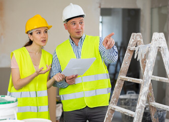 Caucasian man and woman builders discussing project documentation in apartment. Pointing finger gesture.