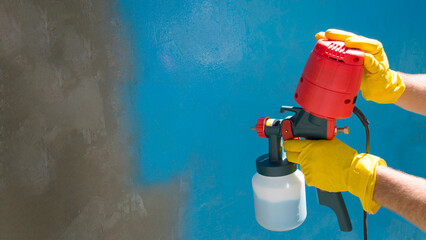 A worker's hands in yellow gloves holds a hand-held spray gun with an overhead compressor. Painting the wall of the house in blue. Household renovation. Copy space. Airless spraying device. Staining