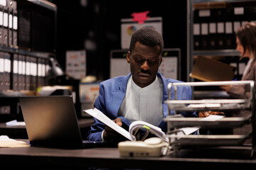 Private detective reading criminology files, searching for new clues to catch mysterious suspect. African american police officer analyzing crime evidence working overtime in arhive room