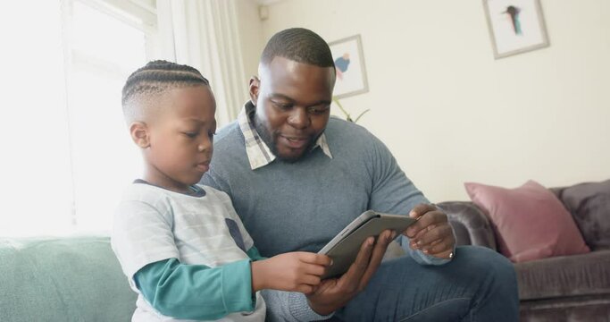 Happy african american father and son sitting on sofa using tablet in living room, in slow motion