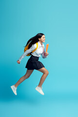 Happy teenage girl wearing uniform, backpack, holding notebooks running fast to school isolated on blue background. Back to school, education concept