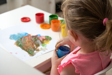 toddler girl sitting at a white table playing with finger paint and holding a can of blue paint in her hands