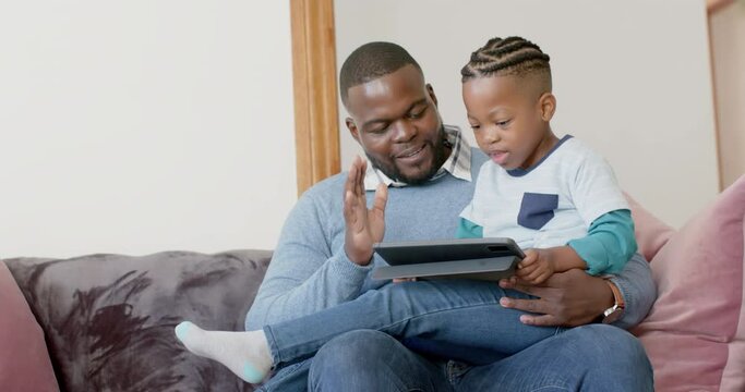 Happy african american father and son sitting on sofa using tablet and high fiving, in slow motion