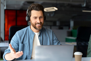 Handsome smiling latin call center operator wearing headphones answering call, communication online sitting in modern office. Successful freelancer working sitting at workplace. Video call concept