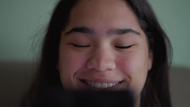 Joyful Brazilian teen girl react's positively to content online while holding phone. Close-up of a happy diverse Asian woman's face smiling reading message