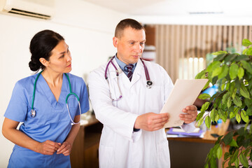 Portrait of two fellow doctors standing in medical office with papers in hands, focused on...