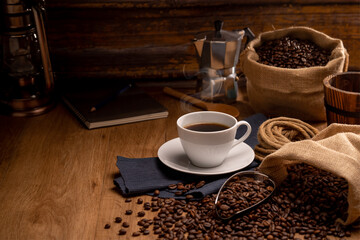 Hot coffee in a white coffee cup and many coffee beans placed around on a wooden table in a warm, light atmosphere, on dark background.