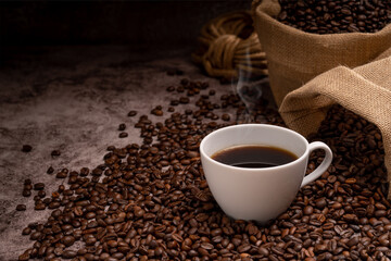 Hot coffee in a white coffee cup and many coffee beans placed around on a wooden table in a warm, light atmosphere, on dark background.