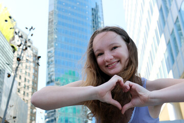 beautiful girl shows heart with hands against backdrop of skyscrapers and big city with huge glass...