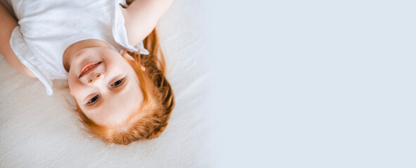 Red-haired girl lies on white bed linen, top view. Charming child with bright hair and freckles.