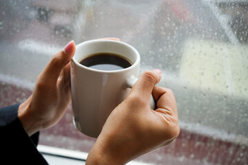 A hand holding a black tablet with a blank white screen on the window with a cup of black latte coffee and sitting on a wooden chair