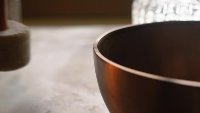 A close-up shot of a mallet striking the rim of a Tibetan singing bowl in slow motion.