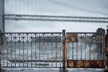 A rusty gate and rusty railing face directly onto a shoreline. Crashing waves and white water are visible in the background The Bay Bridge connecting San Francisco and Oakland is in the distance.