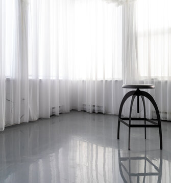 pi&egrave;ce int&eacute;rieur de maison avec des rideaux blancs semi-transparent qui recouvrent les fen&ecirc;tres avec la lumi&egrave;re qui passe au travers ainsi qu'un tabouret en avant plan.