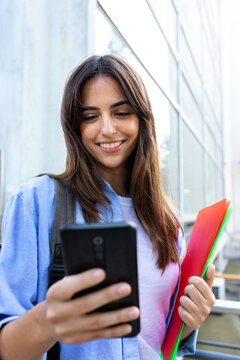 Front view of happy young brunette female university student using smart phone standing outside university. Vertical.