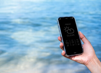 Weather forecast on smartphone. Clear weather, display app on Friday with temperature number, cloud and sun icons on mobile smart phone screen in hand on blue sea water background with copy space.