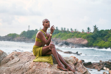 Gay bipoc graceful man poses sitting high on rocks in ocean at sunset. Homosexual slim ethnic...