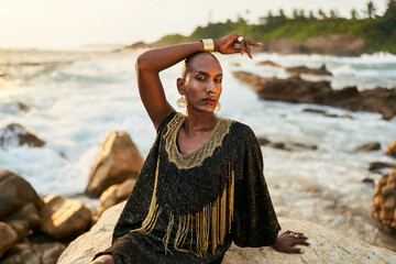 Androgynous ethnic fashion model in luxury dress, jewelry sits on rocks by ocean. Gay black person...