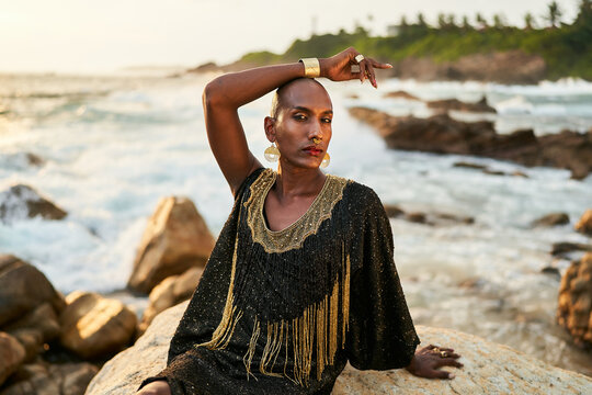 Androgynous ethnic fashion model in luxury dress, jewelry sits on rocks by ocean. Gay black person in jewellery, posh clothes poses gracefully in tropical seaside location. Pride LGBTQ bipoc concept.
