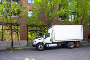 Middle duty day cab rig semi truck with box trailer make delivery standing on the city street with red bricks multilevel apartment building and trees