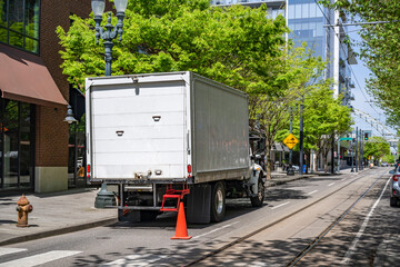 Middle size day cab rig semi truck with box trailer making delivery standing on the city road with warning traffic cone behind the truck