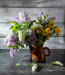 A beautiful still life with lilacs, dandelions, white bird cherry in an old clay teapot on a marble table and a wooden background