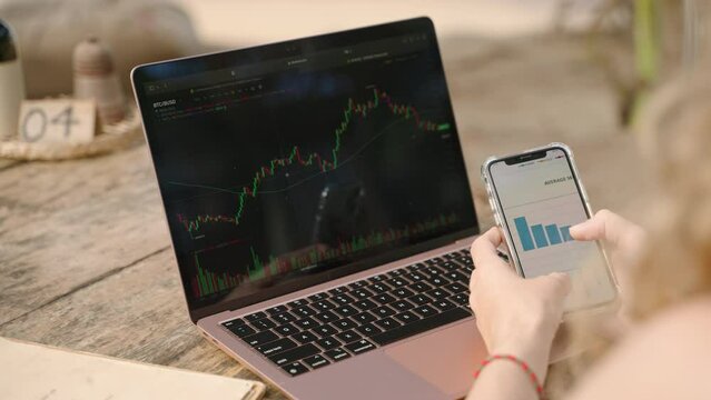 Female crypto trader at laptop with checking statistics charts on smartphone. Woman working remotely with crypto currency checks diagrams on phone. Female analyst studying crypto graphs close-up.