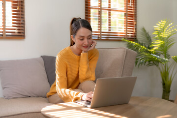 Smiling Asian woman using laptop sitting on sofa at home. Beautiful young Asian female shopping or chatting online in social networks, having fun, watching movie, freelance working on laptop projects.