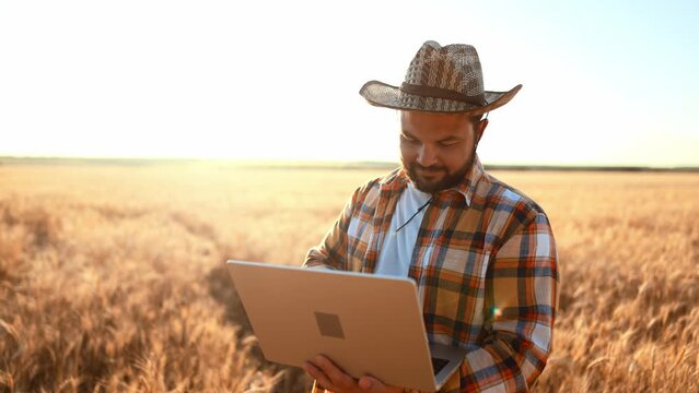 Caucasian farmer with beard in straw hat standing in wheat field and typing on keyboard of laptop computer. Male using device for making profit and improving work in agribusiness and browsing online.