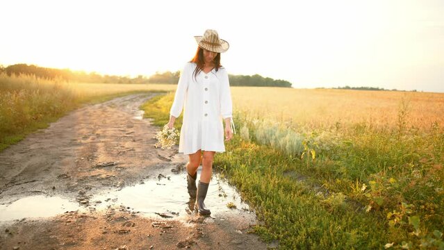 Full length front view of young woman walking along rural road in field with bouquet of chamomiles flowers in her hands. Beautiful girl in straw hat and white dress walks elegantly and looks around.