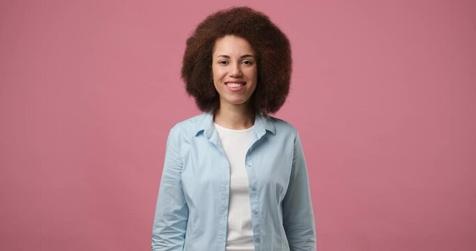 Happy friendly young african american woman waves palm in hello gesture welcomes someone with hospitable expression expresses positive emotions standing over pink background