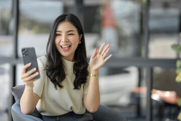 Fototapeta premium Confident young Asian woman using mobile phone video call with co-workers showing joy and admiration for successful online business together while sitting on sofa chair at office.