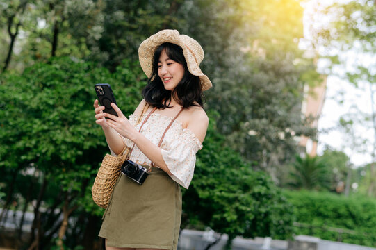Portrait of asian young woman traveler with weaving hat, basket, mobile phone and camera on green public park background. Journey trip lifestyle, world travel explorer or Asia summer tourism concept.