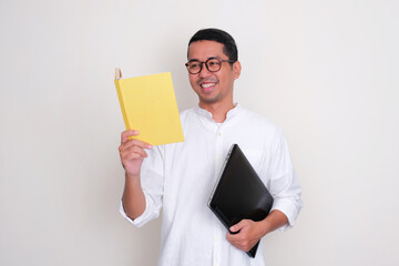Adult Asian man smiling happy when reading a book and holding laptop