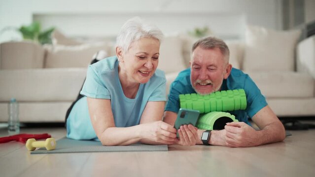 Senior couple man and woman watching video on smartphone after home sport training lying on mats on floor in living room. Laughing, talking discussing watched in phone having rest, relaxing together.