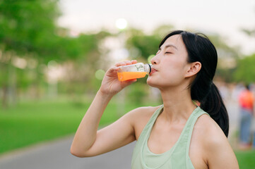 Female jogger. Fit young Asian woman with green sportswear drinking organic orange juice after running and enjoying a healthy outdoor. Fitness runner girl in public park. Wellness being concept
