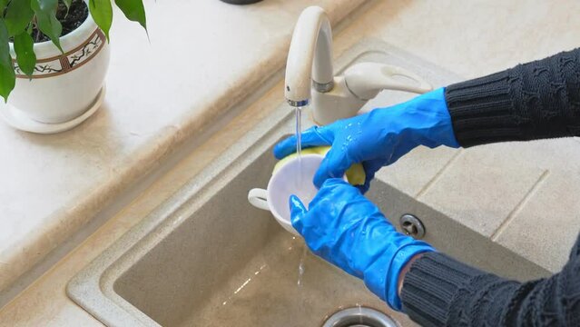 Maid use sponger wear blue latex gloves washes dishes in the ceramic sink on house kitchen indoors . A domestic worker woman wash coffee cups after guests arrive. Housewife girl doing household chores