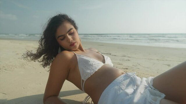 Medium view of tan indian woman in bikini looking up and out as she lays on sandy beach